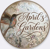 Round decorative sign featuring a painted hummingbird on the left and the words 'April's Gardens' in dark brown cursive script over a soft floral background.